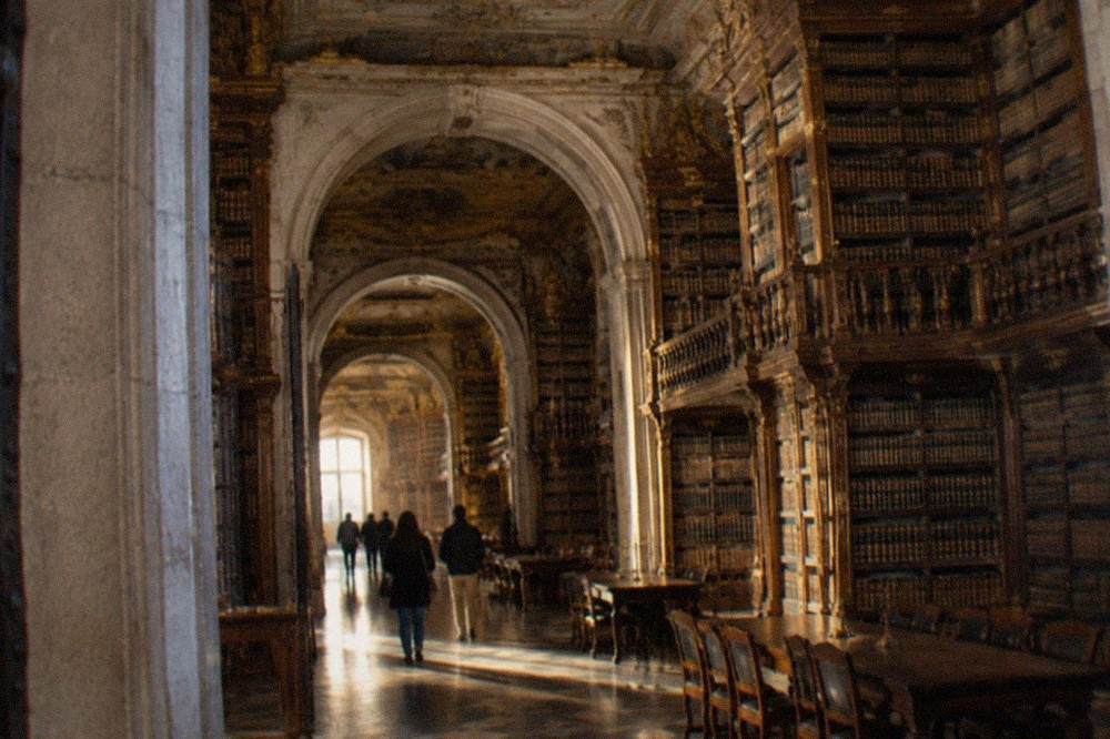Visitors walking through the arched stone entrance of the Joanina Library building at the University of Coimbra