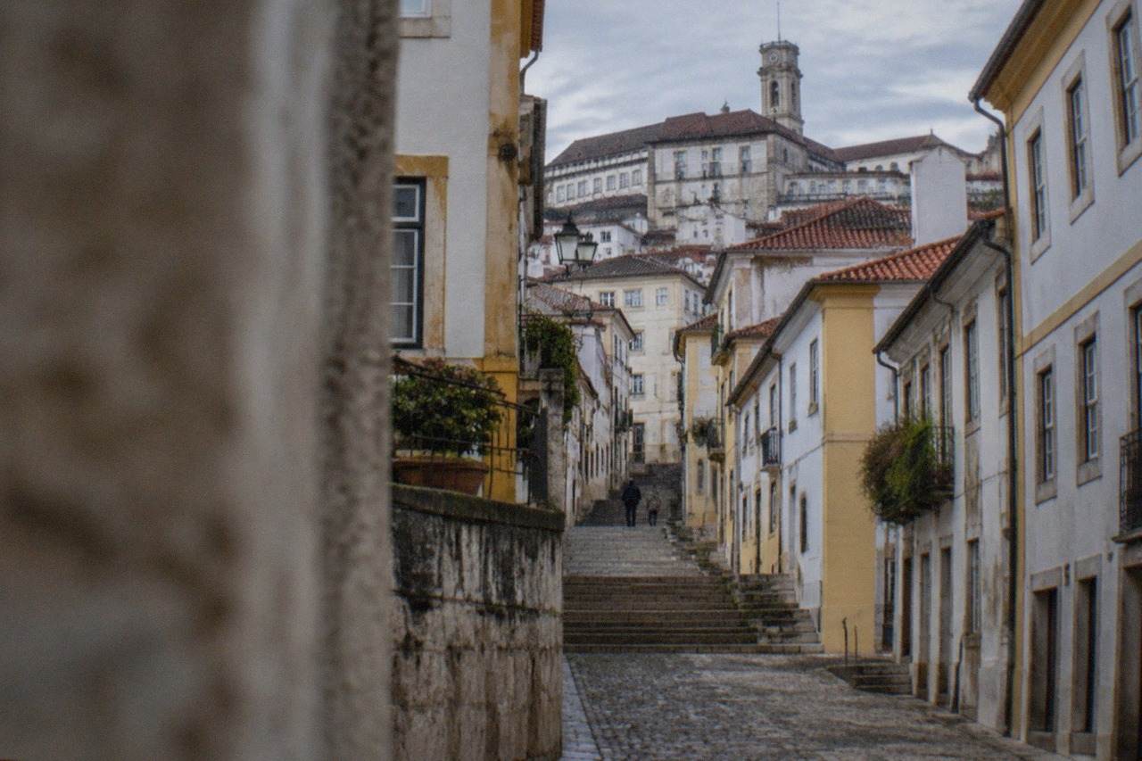 A cobbled street rising toward the university buildings of Coimbra, Portugal, with stone steps and old houses on either side.