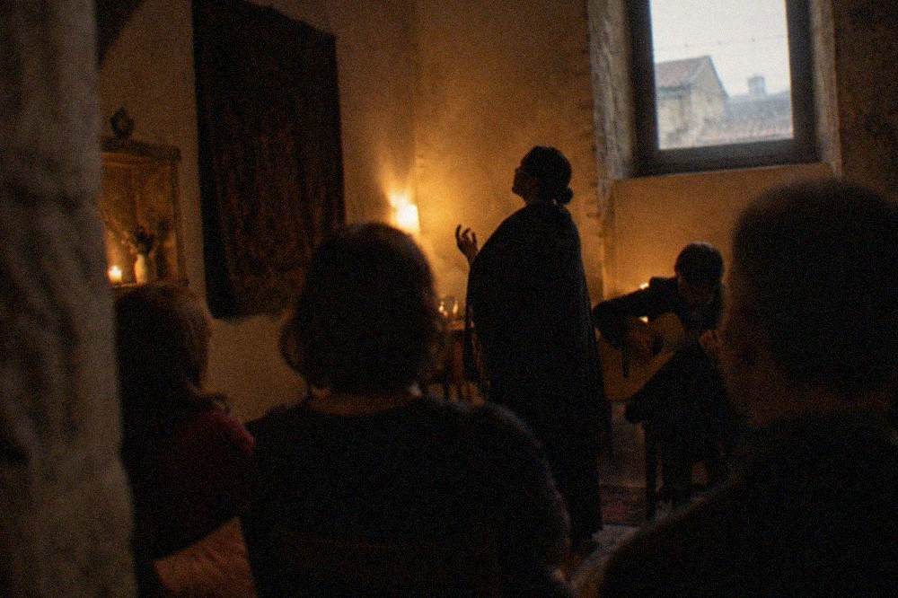 A dimly lit room where a singer in a dark academic cloak performs fado to a quiet audience