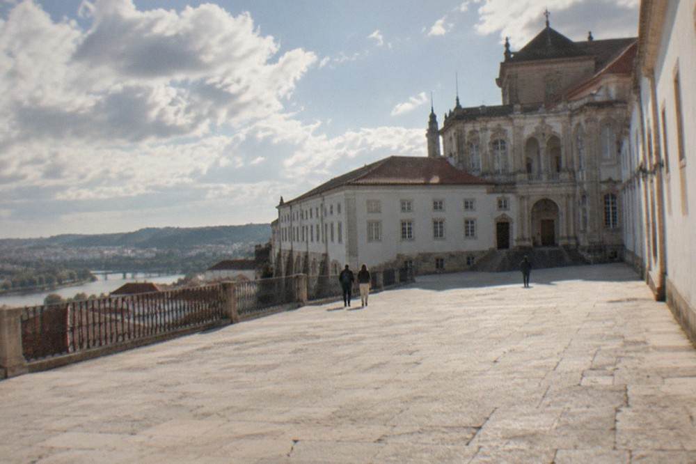 The lit facade of the University of Coimbra's Paço das Escolas at dusk seen from a narrow street below