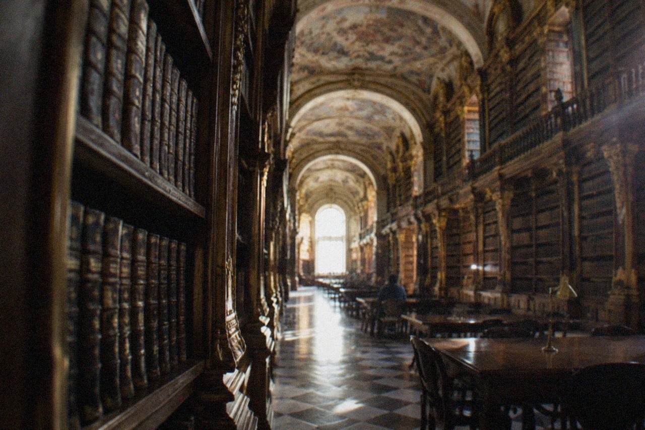 The ornate baroque interior of the Joanina Library at the University of Coimbra, with tall wooden bookshelves and painted ceilings