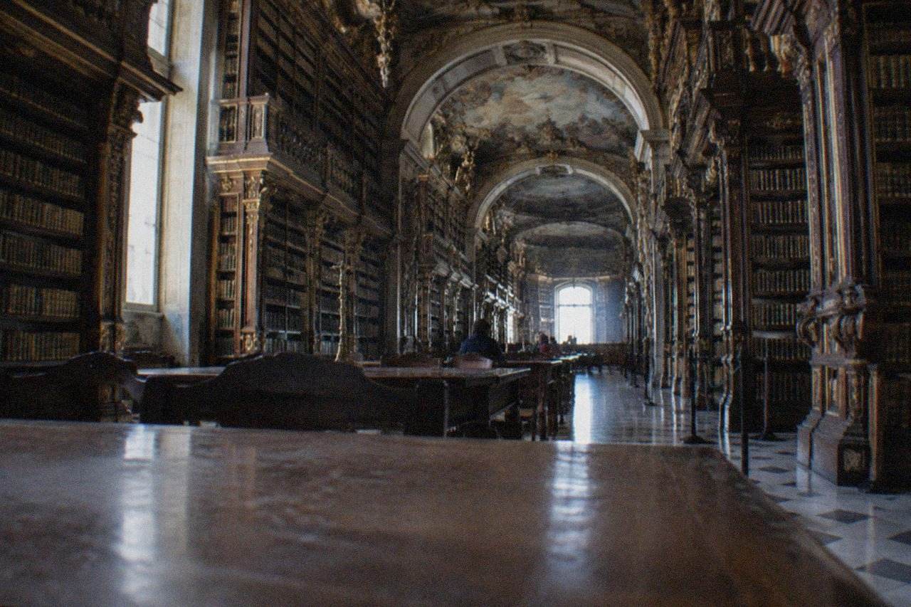 The ornate baroque reading room of the Joanina Library in Coimbra, with tall wooden bookshelves reaching to a painted ceiling
