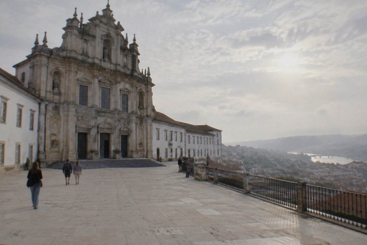The grand baroque facade of the University of Coimbra seen from below, with the city's rooftops and the River Mondego in the background