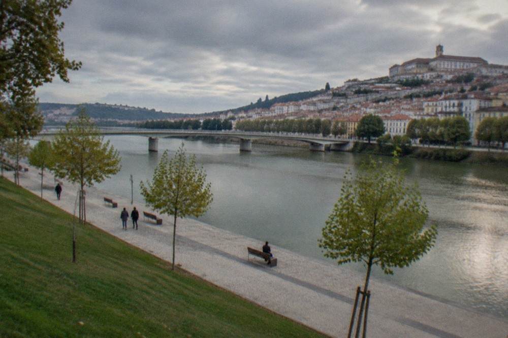 A wide paved riverside walk along the Mondego with trees, benches and the bridge to the old town in the distance