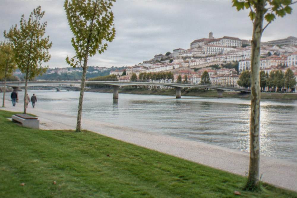 A riverside lawn beside the Mondego with the flat Santa Clara ground and the hillside city rising across the water