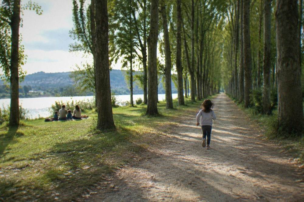 A family having a picnic on grass under tall trees near the riverbank