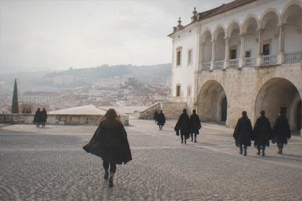 Students in black capes walking across a cobbled square in front of a grand university building