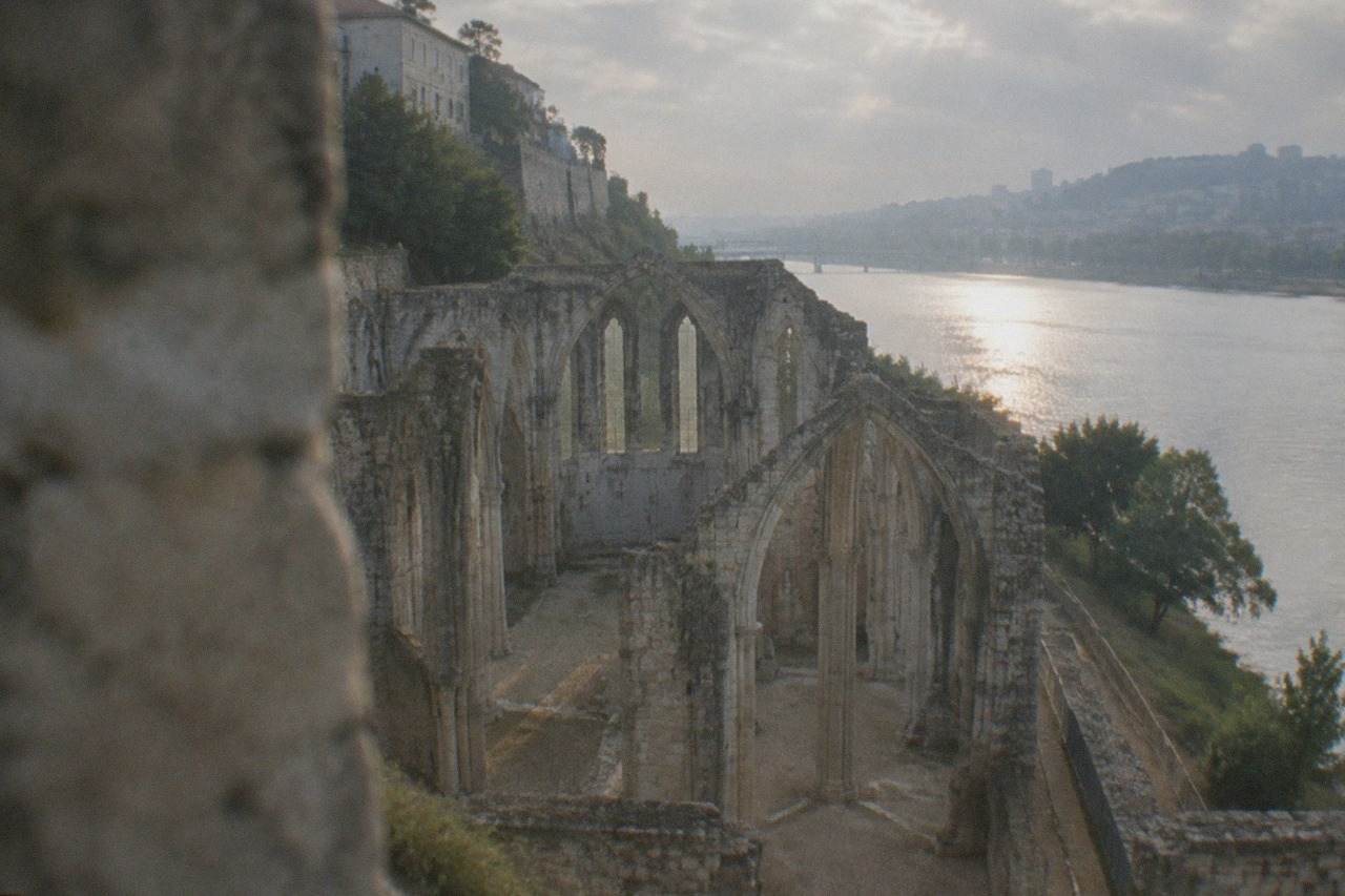 Stone ruins of an old monastery church beside a river, with the walls of a later building on the hill above