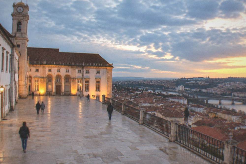The lit facade of the University of Coimbra at dusk with the city rooftops of Coimbra below