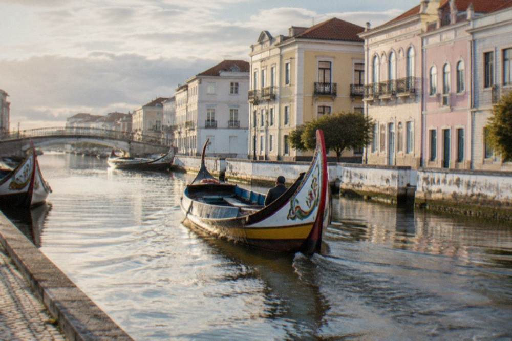 A moliceiro boat gliding along a narrow canal between low pastel buildings in Aveiro