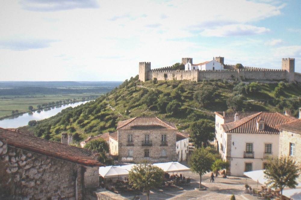 A small Portuguese town square with stone buildings and café tables below a castle hill