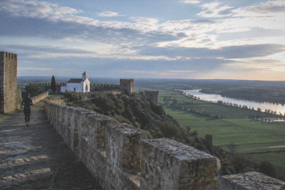A view across crenellated castle walls toward a wide green river plain under a big sky