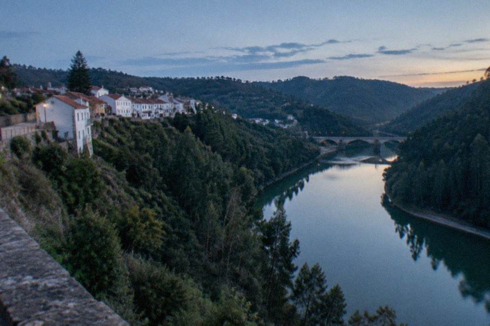 A calm river bend reflecting trees, with a stone bridge in the distance under a clear sky