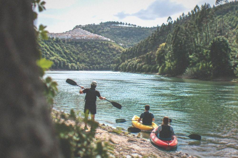 A guide demonstrating paddle strokes to two seated kayakers near a riverbank