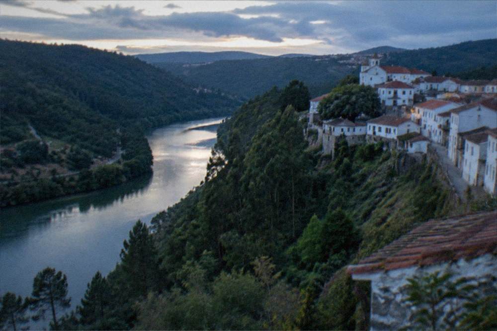 A hillside stone village among trees seen across a river valley in late afternoon light