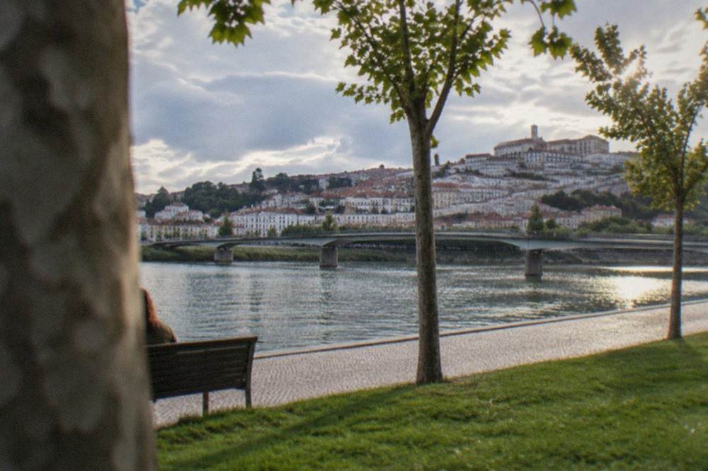 A level riverside walkway along the Mondego with the old bridge and city skyline behind it