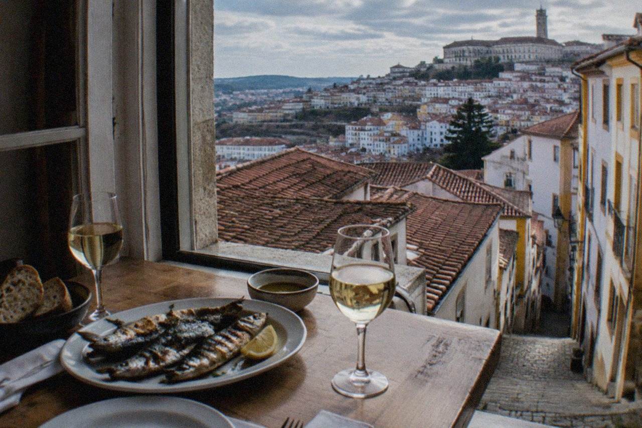A laid table with grilled fish, bread and wine beside a window overlooking tiled Coimbra rooftops