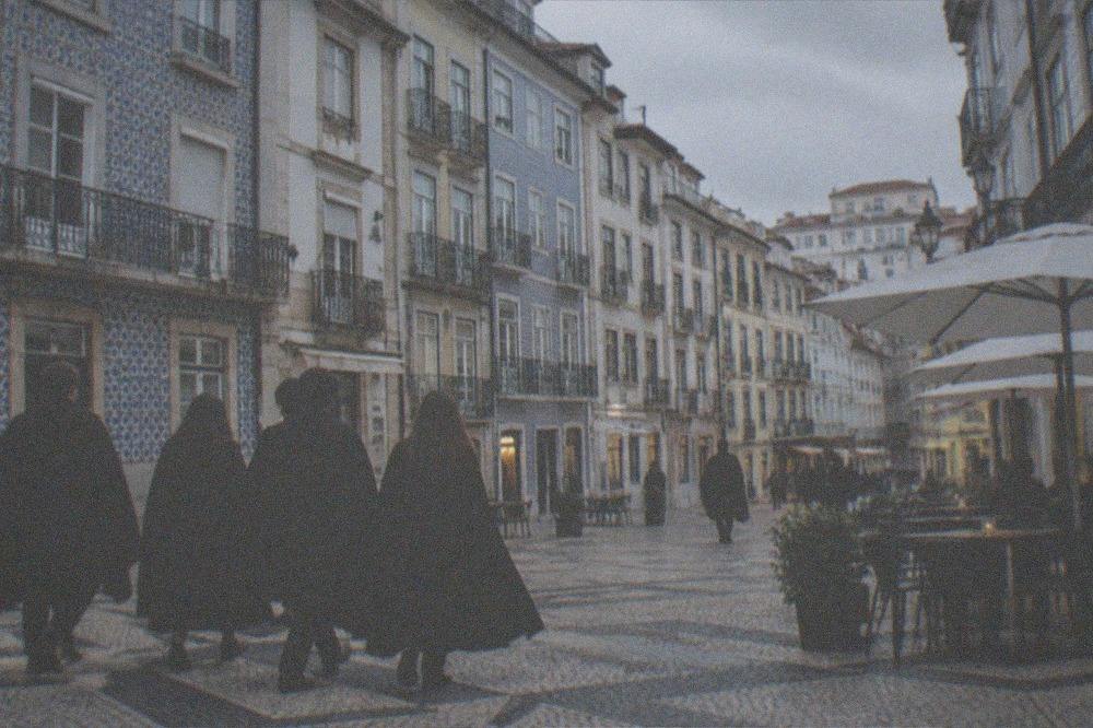 Students in black capes crossing a cobbled square past café tables in the evening