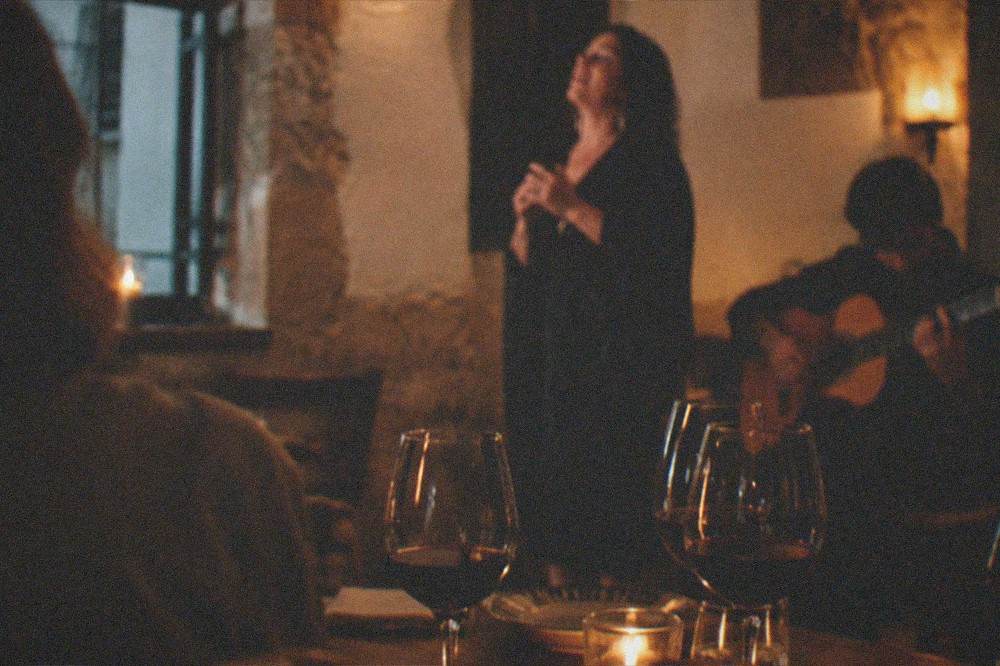 A dinner table with wine glasses in a dim room while a singer performs fado nearby