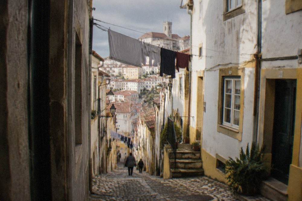 A shaded cobbled street in Coimbra's old town with sunlit walls and laundry overhead