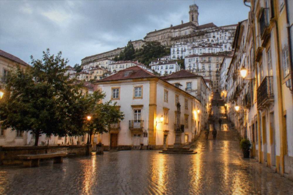 A rainy evening in a Coimbra square with wet cobbles reflecting warm street lights
