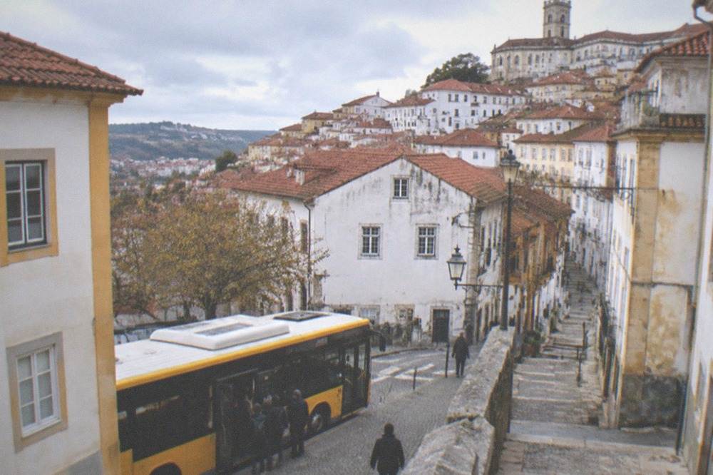 A yellow city bus stopped at a street in central Coimbra with passengers boarding