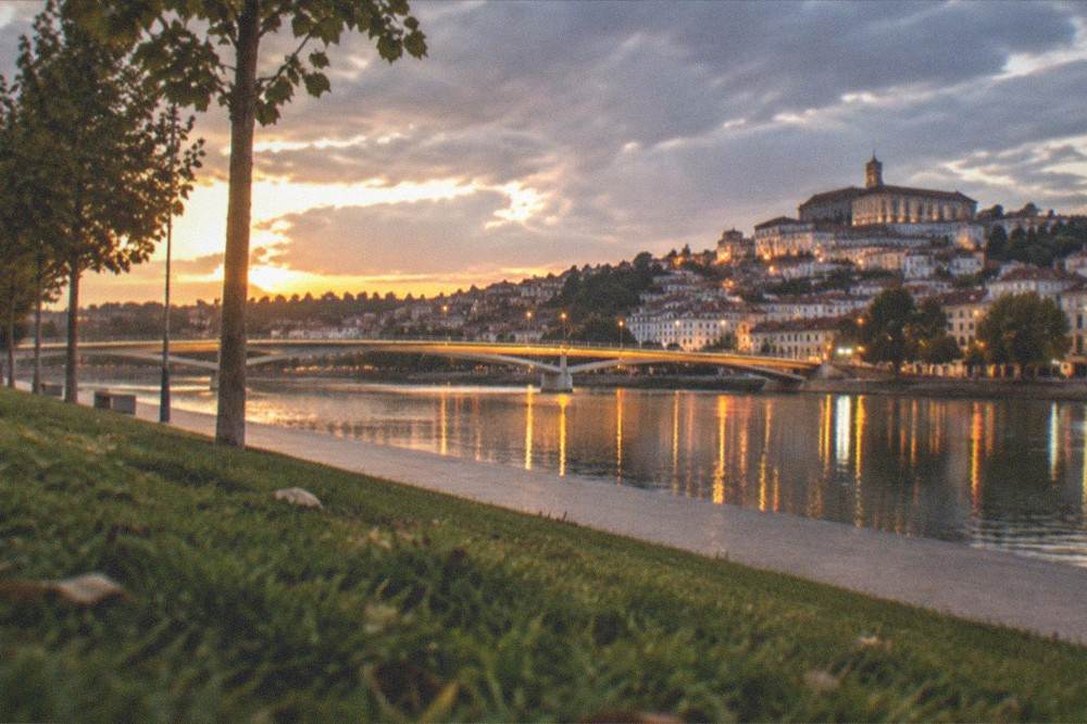 An evening view of an illuminated bridge over the Mondego river with the city lights of Coimbra reflected in the water