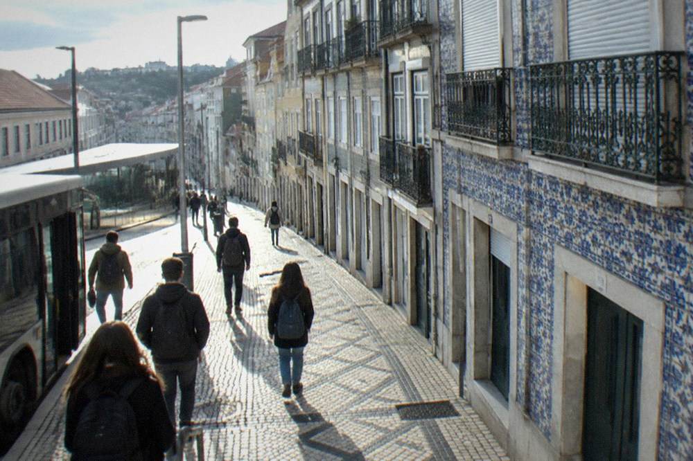 Passengers with backpacks walking out of a bus terminal onto a city street lined with low buildings