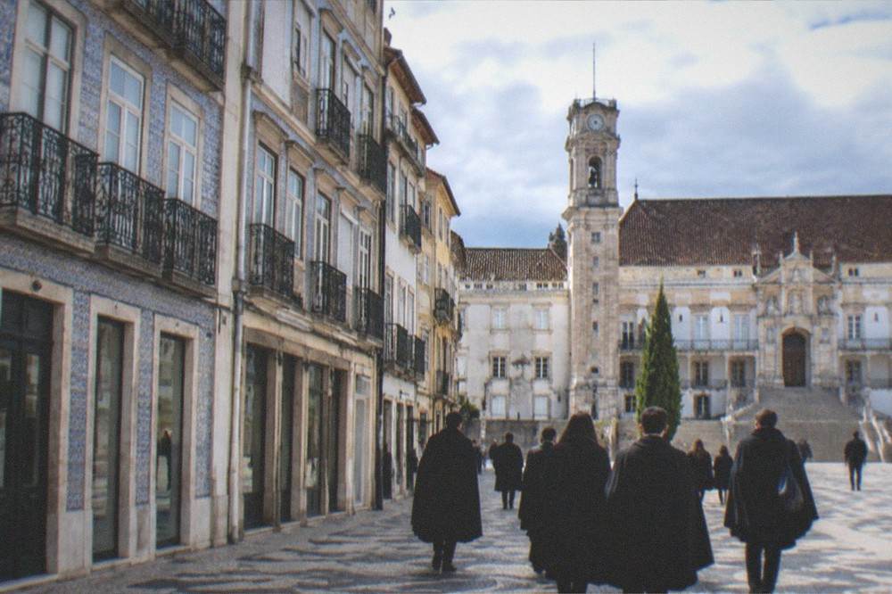 Students in black capes crossing a cobbled square in front of the university buildings