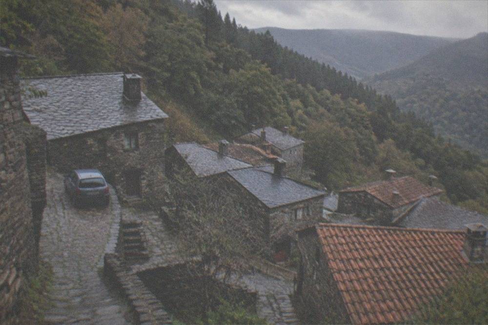 A small car parked on a stone road beside a schist village house on a forested mountain slope
