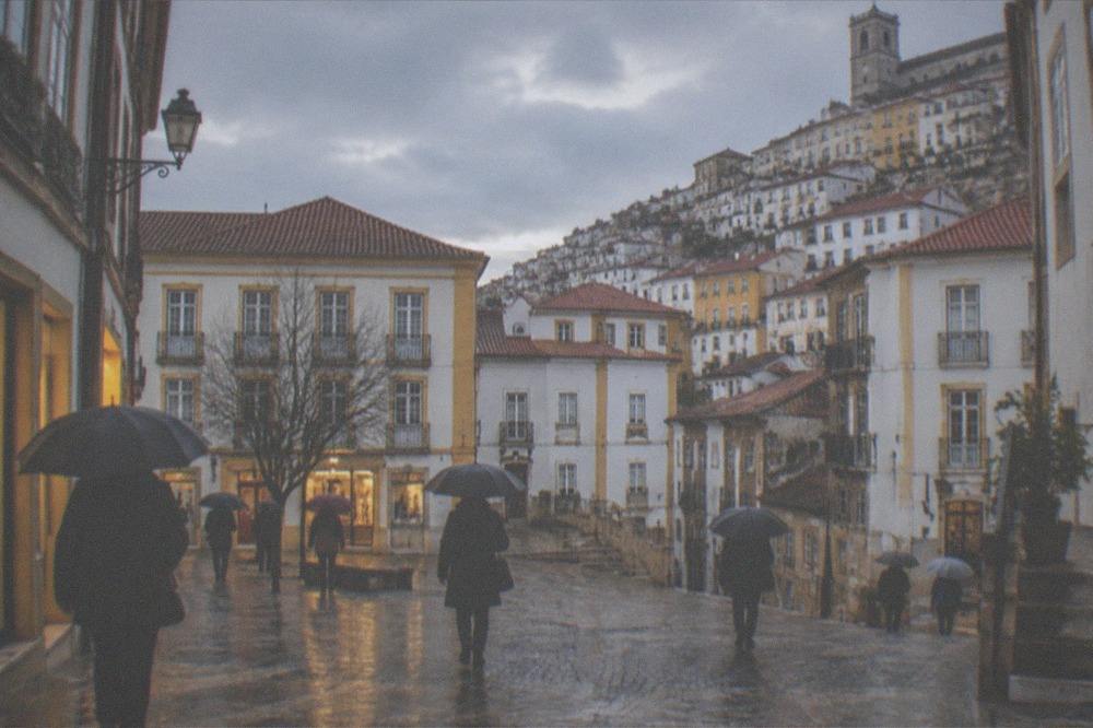 Rain-slicked cobbled square in Coimbra with people under umbrellas walking past lit shopfronts