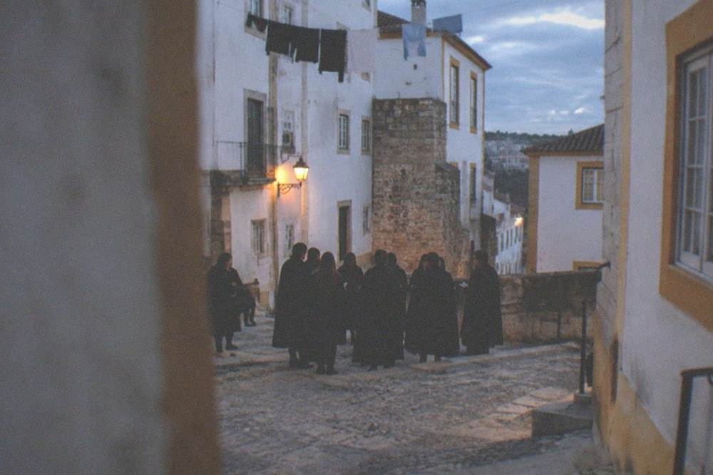 A group of students in black capes singing together in a stone square at dusk