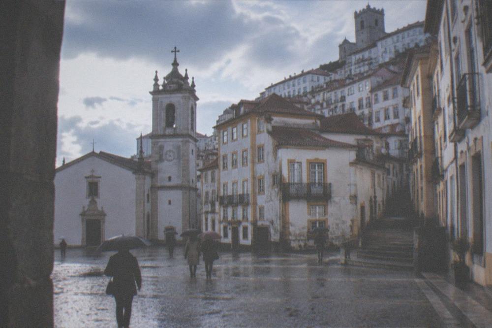 Visitors with umbrellas walking across a wet cobblestone square with a church tower behind