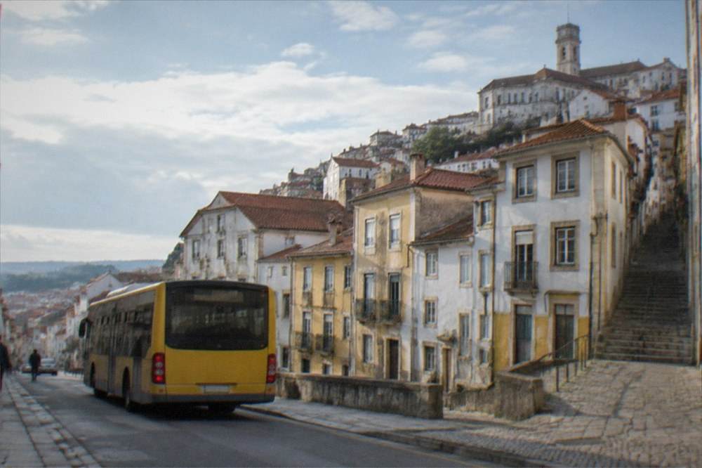 A yellow city bus stopped on a street with a steep hill and old buildings