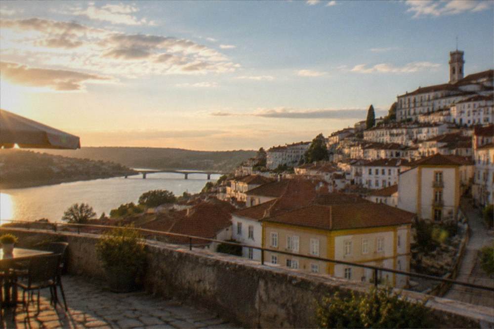 Sunlit terrace overlooking the tiled roofs of Coimbra with the river valley beyond in warm evening light.