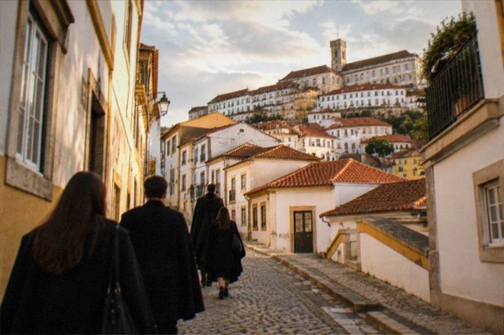 Students in black capes walking up a cobbled street toward the university hill in Coimbra