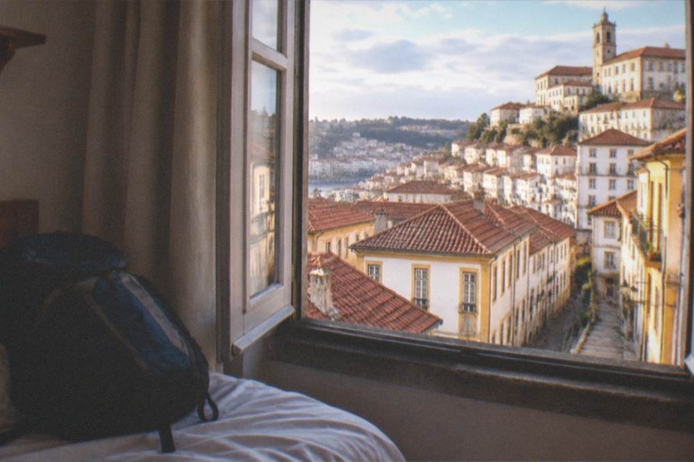 Backpack resting on a hostel bed beside a window with Coimbra rooftops beyond