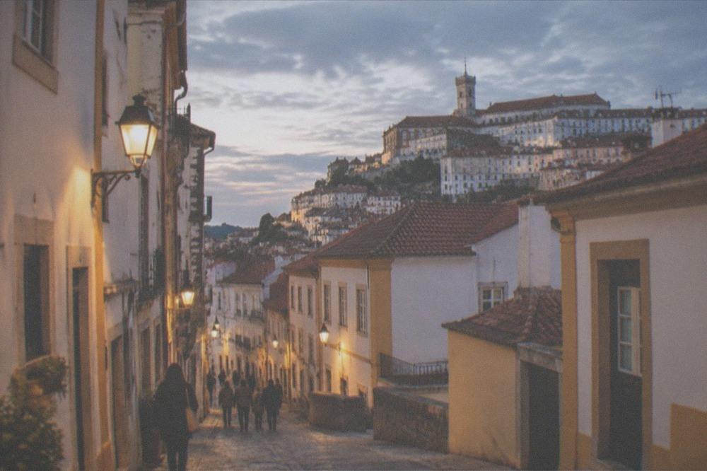 An evening street in a Portuguese old town lit by warm lamps with people walking