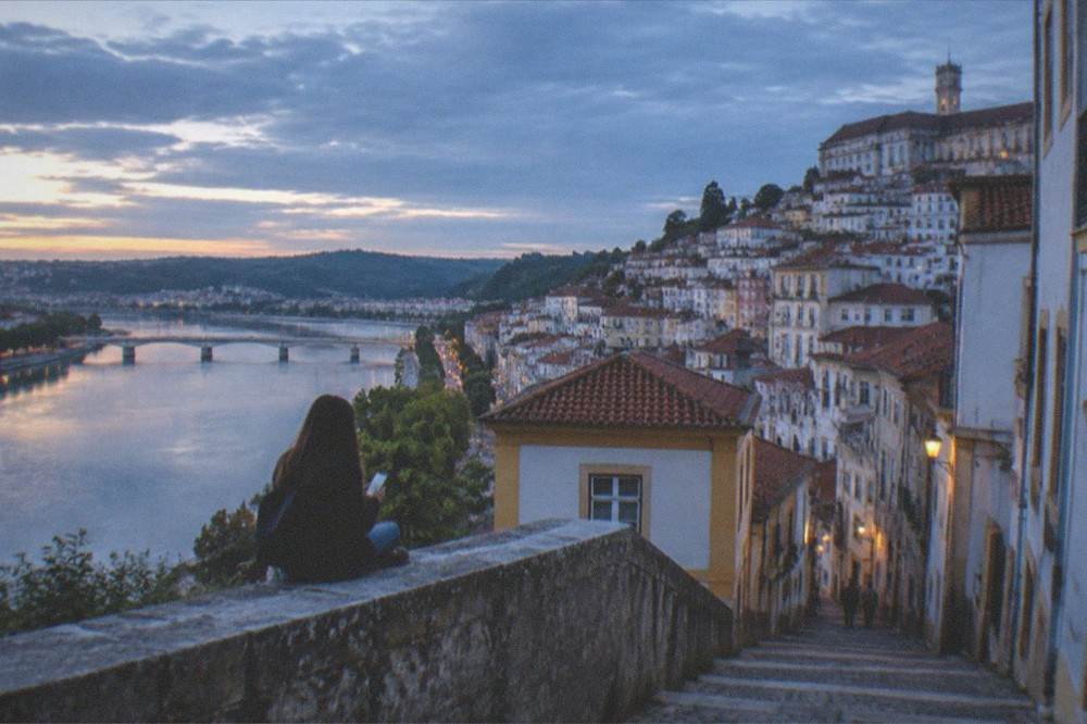 Traveller checking a phone while sitting on a wall overlooking the city of Coimbra and its river