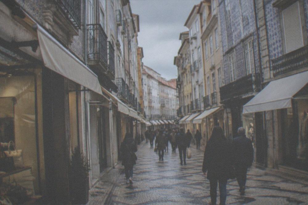 A narrow shopping street in the Baixa of Coimbra with awnings and shoppers in the late morning