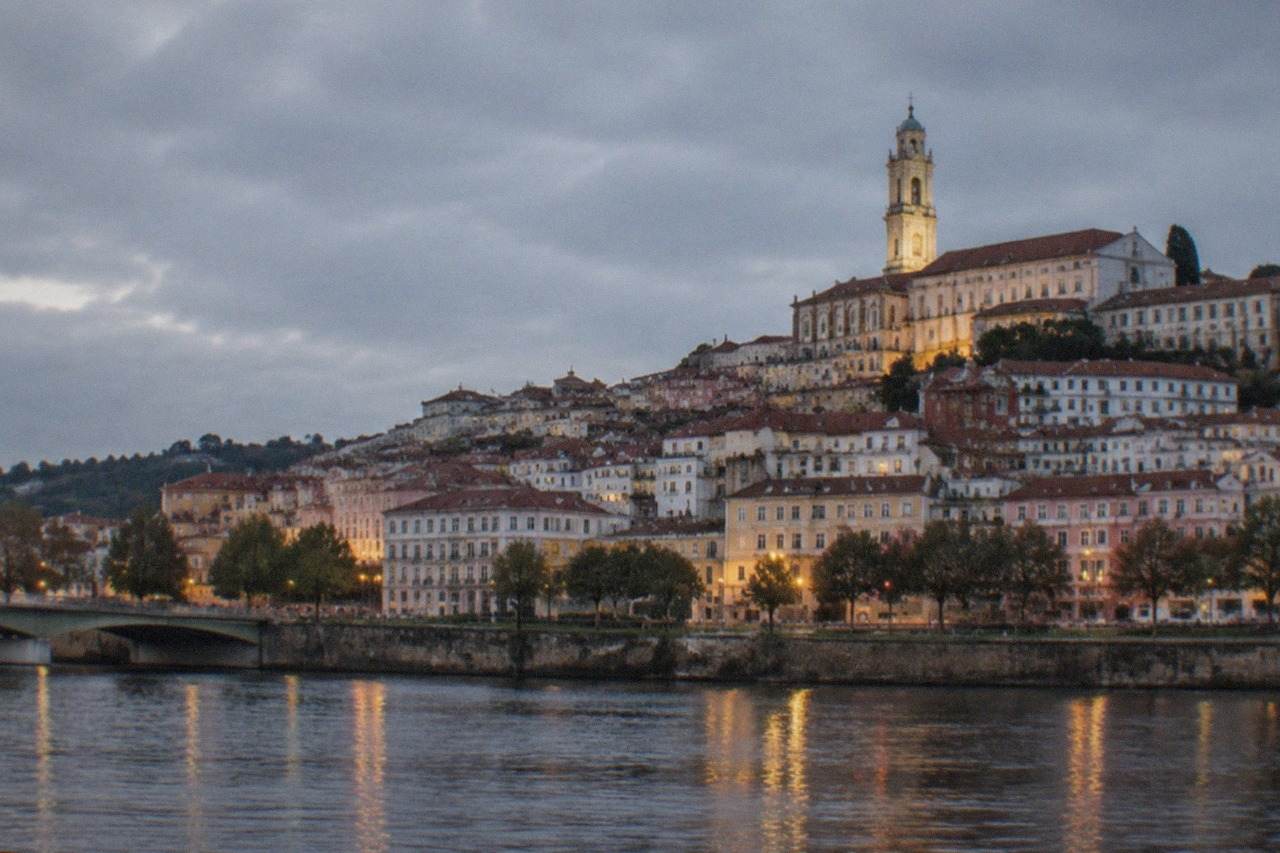 The illuminated old town of Coimbra seen at dusk from across the River Mondego, with the university tower lit above the rooftops