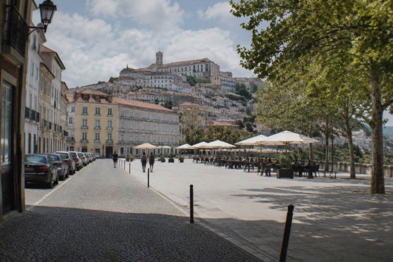 A narrow cobbled street in Coimbra's old town with parked cars lining one side and the university hill rising behind