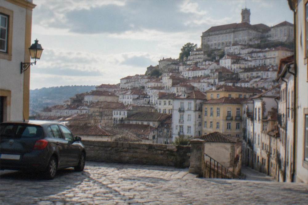 A parked car at the edge of a flat square in Coimbra with the old town hill in the background