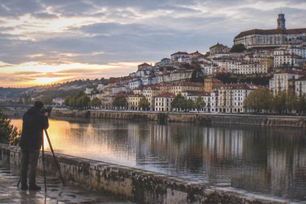 A photographer with a camera and tripod photographing the city of Coimbra from a riverside viewpoint at dusk