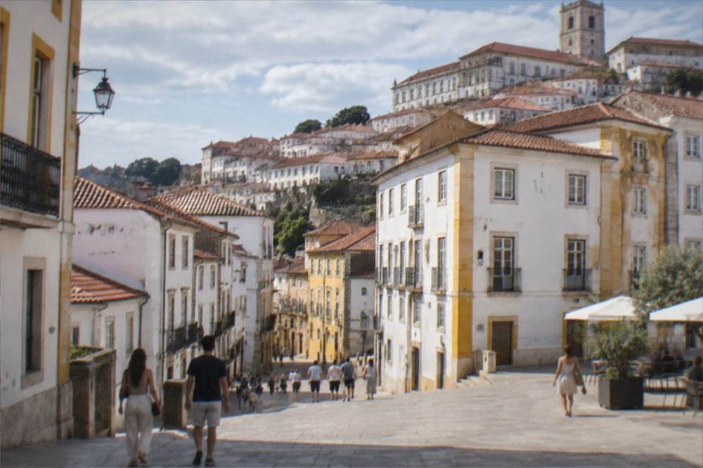 Visitors in light clothing walking across a sunlit square in a Portuguese university city