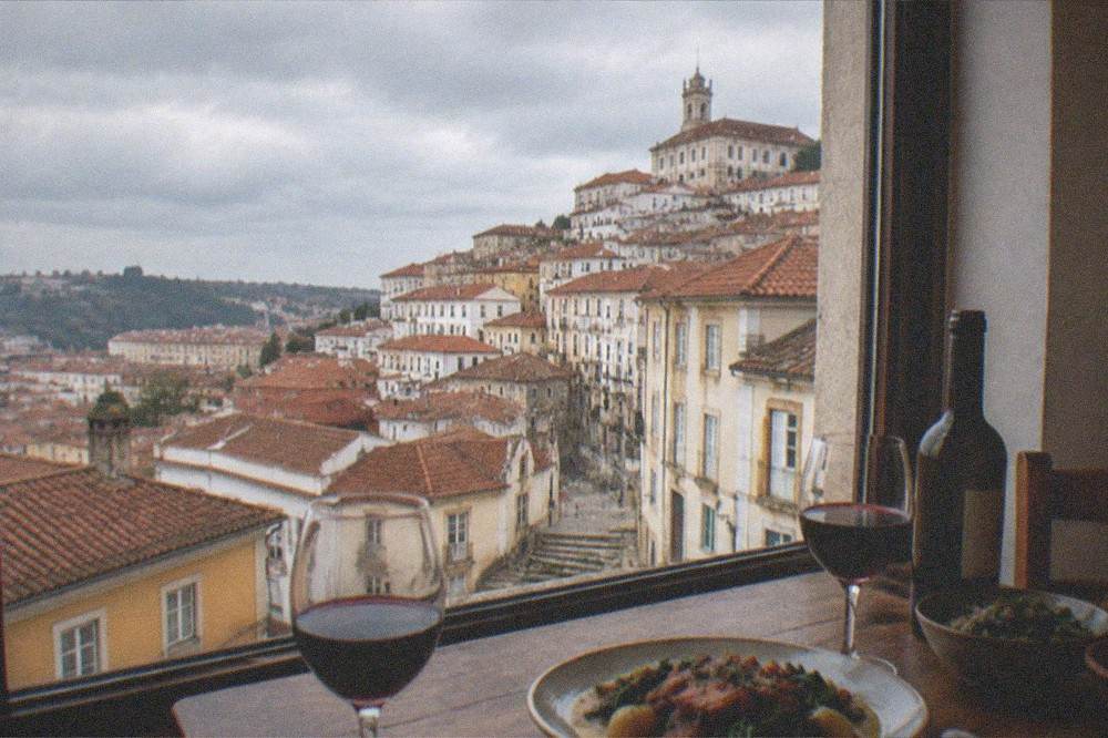 A dining table with a bottle of red wine, glasses and a plate of regional Portuguese food beside a window overlooking Coimbra rooftops