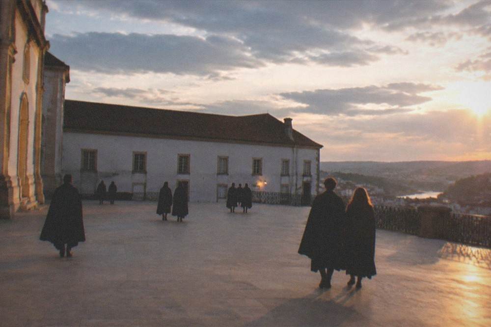 The illuminated courtyard of the Paço das Escolas at night with students in black capes crossing it