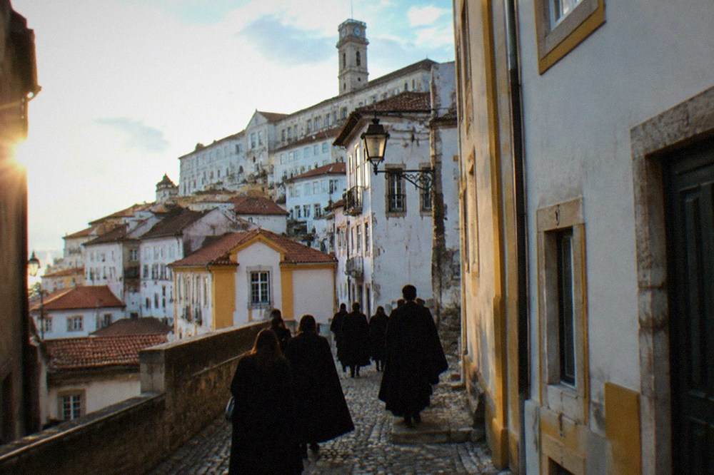Students in dark capes walking up a cobbled street toward the university buildings in Coimbra.