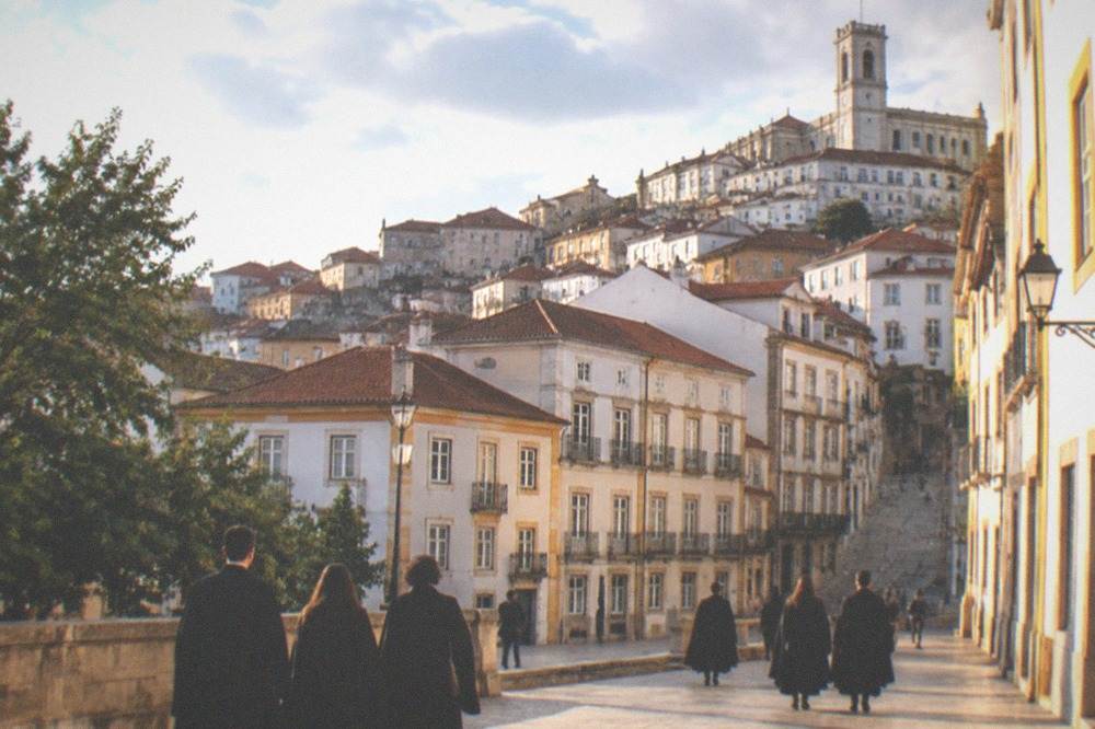 Students in black capes walking through a Coimbra square on a warm evening