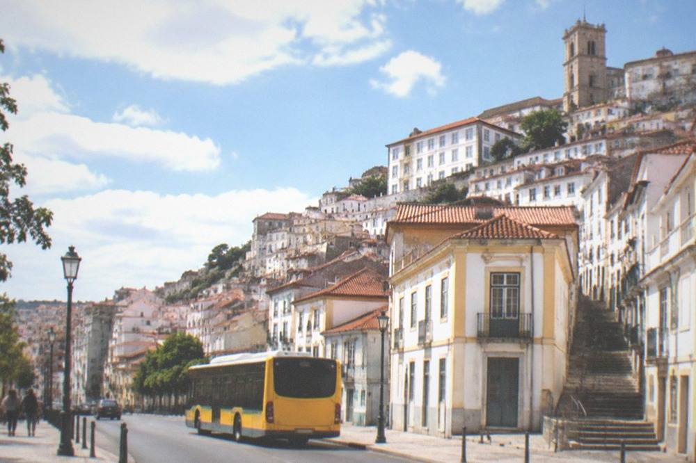 A yellow city bus on a street in central Coimbra with the old town rising behind it